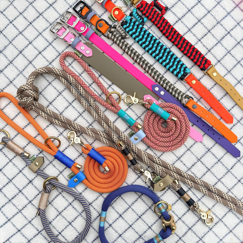 Flat lay of handmade custom dog accessories on blue checked fabric: rope leads in orange, navy & chestnut, paracord collars with BioThane straps, and a marine blue yarn collar. Brass, nickel & black hardware. Fully customisable.