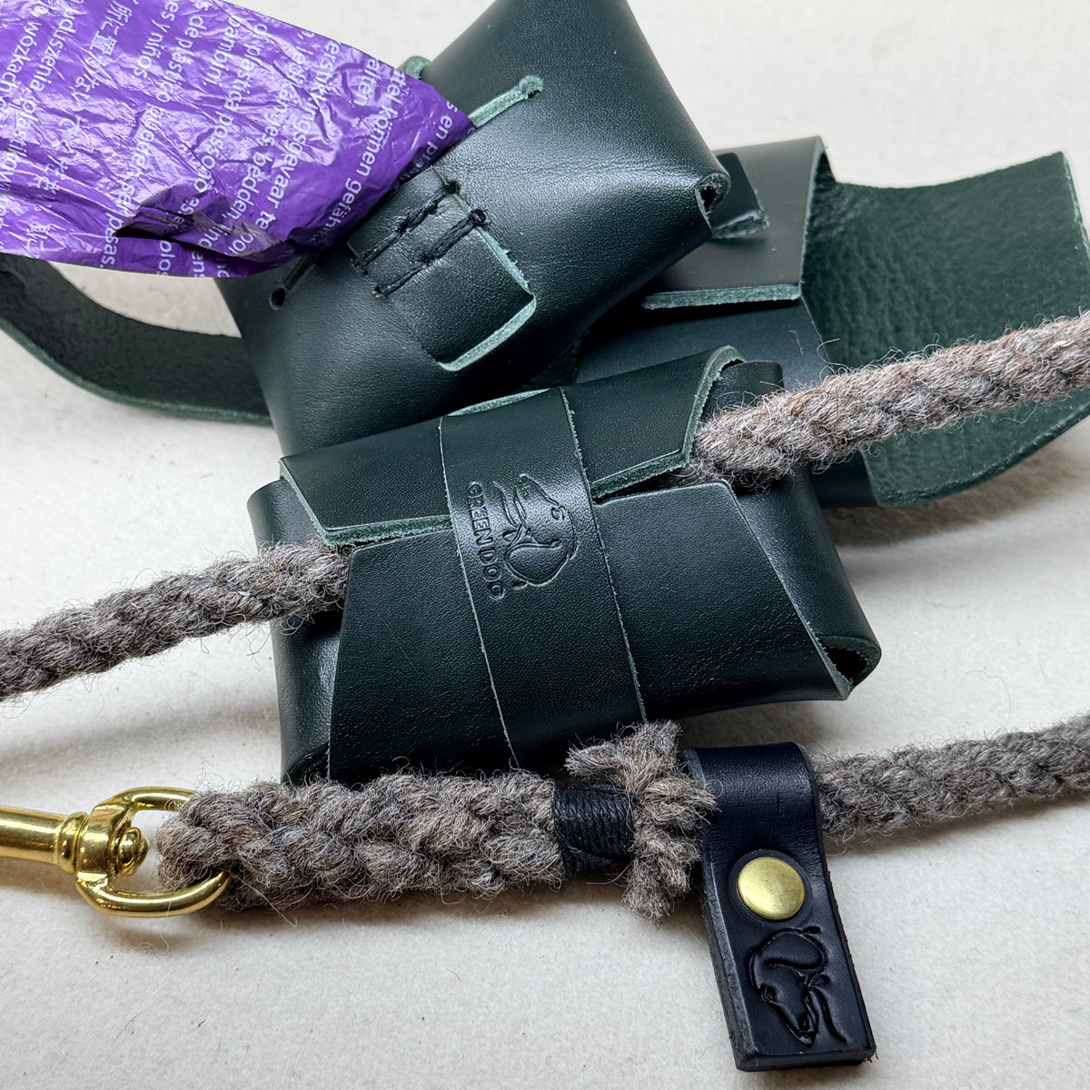 Hunter green leather poo bag holder with a hand stitched saddle stitch seam, wrapped around a wool rope dog lead with a purple compostable poo bag tucked inside
