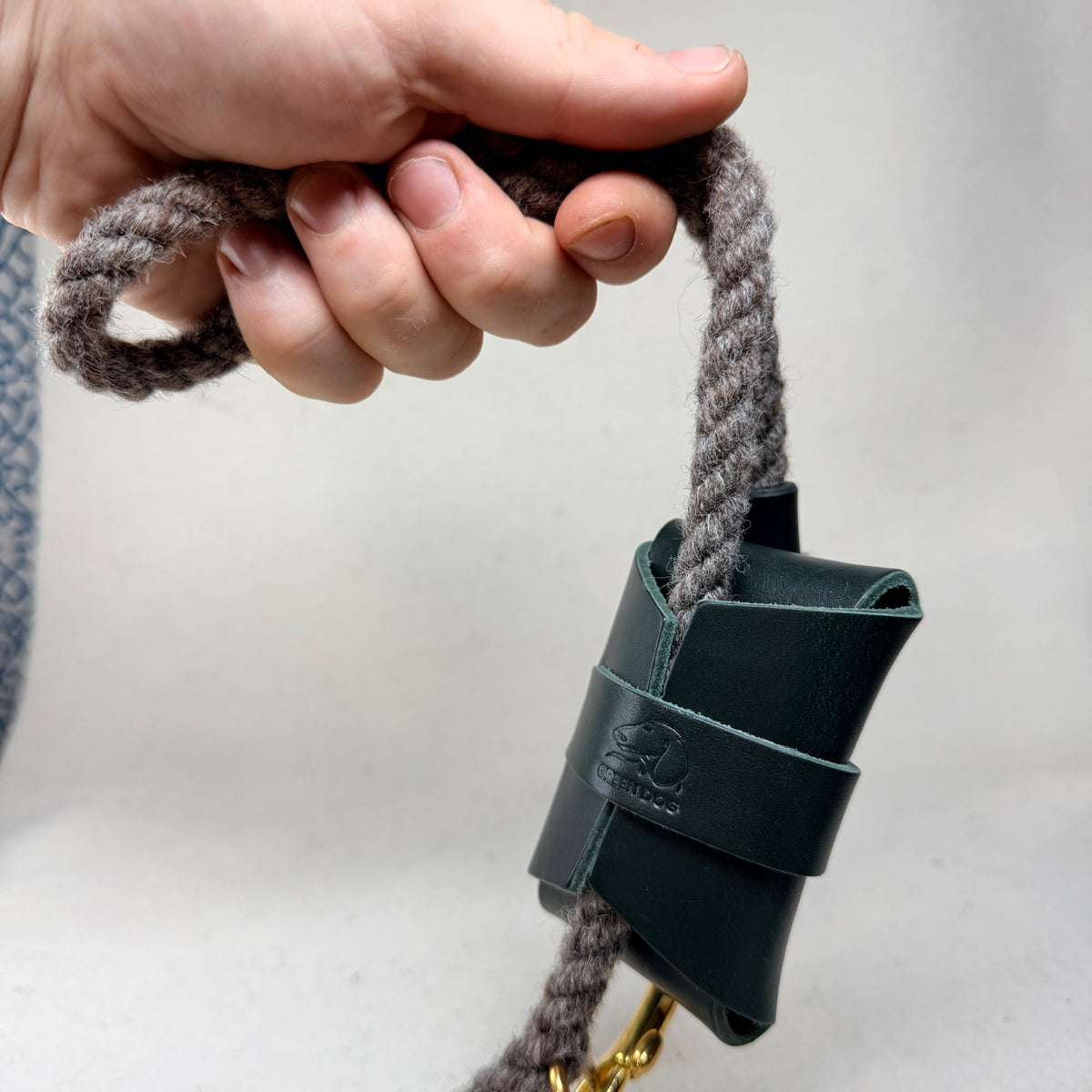 Hunter green leather poo bag holder hanging from a wool rope dog lead held in hand, showing the hand embossed Green Dog logo